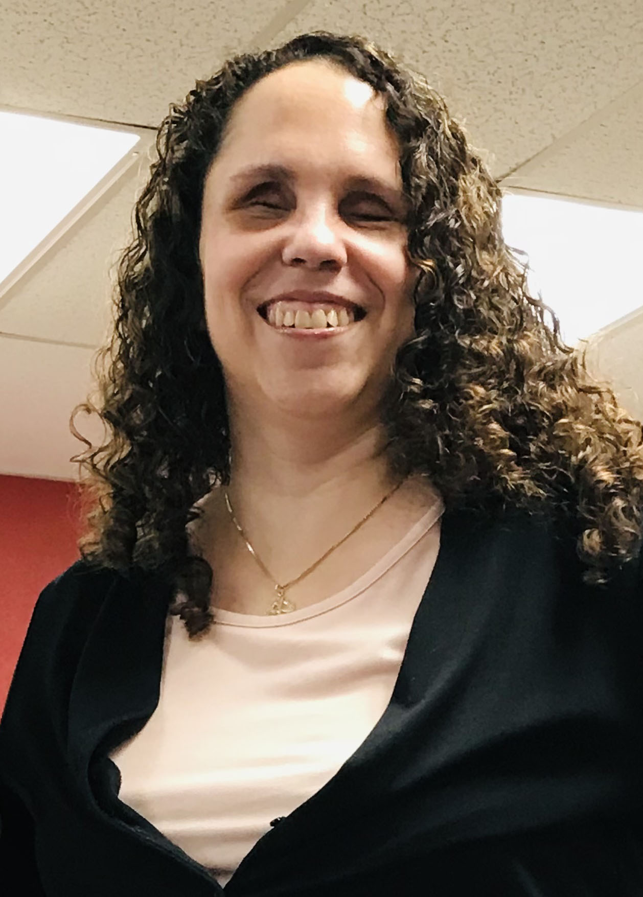 A close up of Julia smiling with long brown curly hair, wearing a black sweater and light pink top