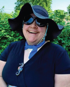 Kirstianna smiling and wearing a large brimmed black hat, dark glasses, and a black dress with seahorse brooch